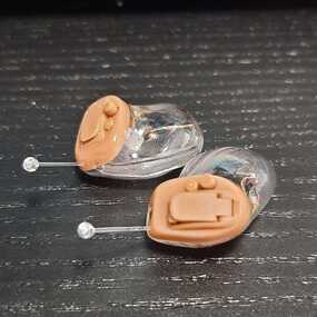 Zwei moderne, transparente In-Ear-Hörgeräte mit Elektronik auf schwarzer Oberfläche.