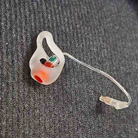 Transparenter Ohrpassstück mit Filter für Hörgerät liegt auf dunklem Stoffhintergrund.