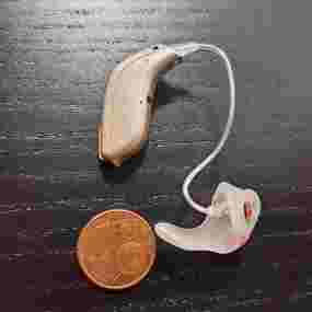 Modernes Hörgerät mit Ohrpassstück neben einer Münze auf dunklem Holztisch.